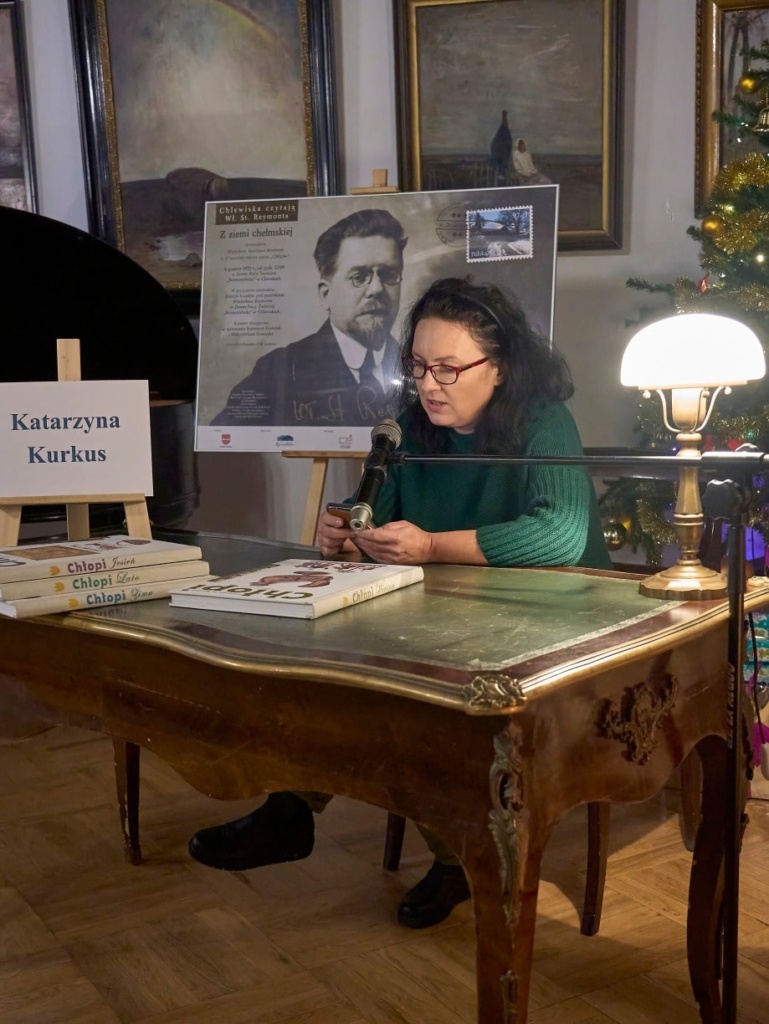 Kobieta okularach siedzi przy zabytkowym biurku, czytając tekst do mikrofonu podczas wydarzenia literackiego. Na stole przed nią znajdują się książki z serii „Chłopi” Władysława Reymonta. Obok stoi tabliczka z jej imieniem i nazwiskiem: „Milena Jastrzębska”. W tle widoczny jest plakat z portretem Władysława Reymonta oraz udekorowana choinka z ozdobami.