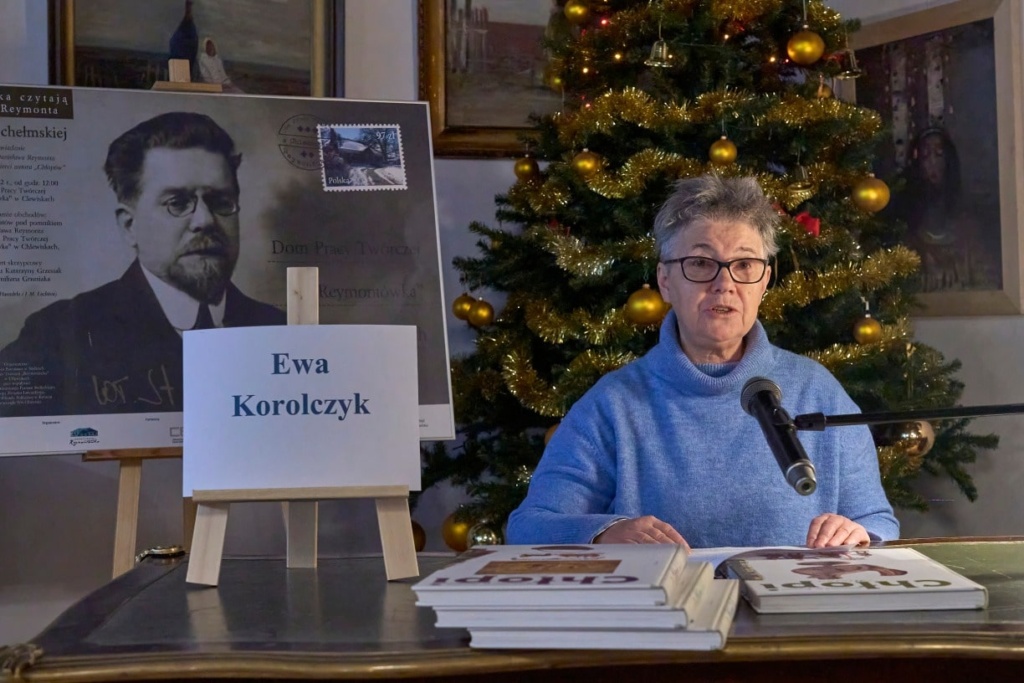 Kobieta w okularach siedzi przy zabytkowym biurku, czytając tekst do mikrofonu podczas wydarzenia literackiego. Na stole przed nią znajdują się książki z serii „Chłopi” Władysława Reymonta. Obok stoi tabliczka z jej imieniem i nazwiskiem: „Milena Jastrzębska”. W tle widoczny jest plakat z portretem Władysława Reymonta oraz udekorowana choinka z ozdobami.