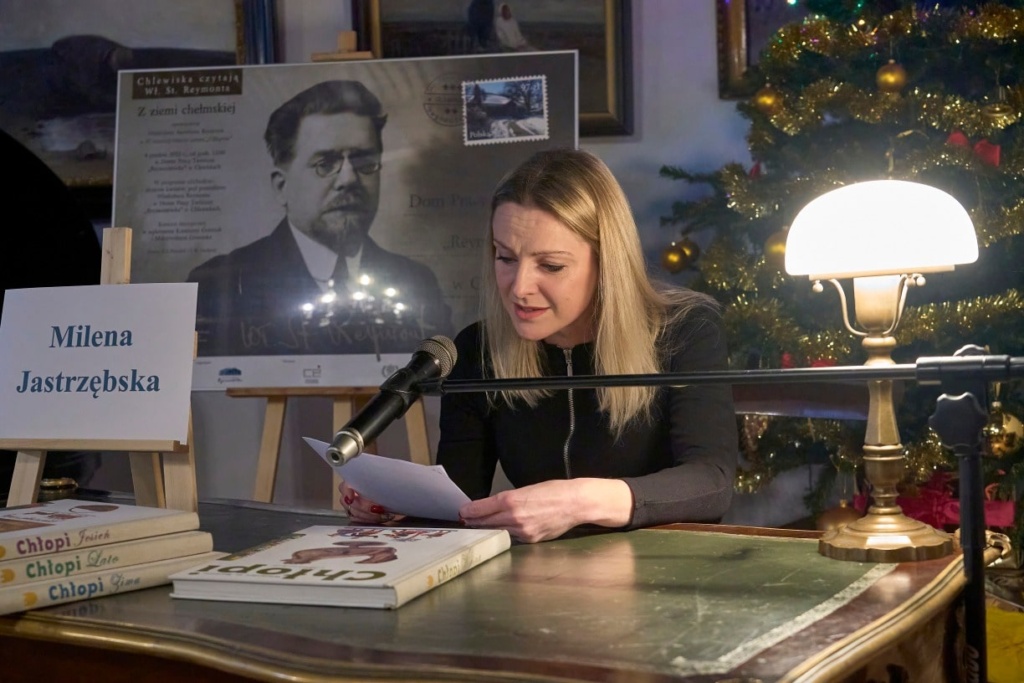 Kobieta o blond włosach siedzi przy zabytkowym biurku, czytając tekst do mikrofonu podczas wydarzenia literackiego. Na stole przed nią znajdują się książki z serii „Chłopi” Władysława Reymonta. Obok stoi tabliczka z jej imieniem i nazwiskiem: „Milena Jastrzębska”. W tle widoczny jest plakat z portretem Władysława Reymonta oraz udekorowana choinka z ozdobami.