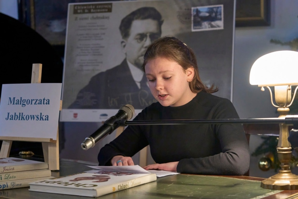 Kobieta o brunatnych włosach siedzi przy zabytkowym biurku, czytając tekst do mikrofonu podczas wydarzenia literackiego. Na stole przed nią znajdują się książki z serii „Chłopi” Władysława Reymonta. Obok stoi tabliczka z jej imieniem i nazwiskiem: „Milena Jastrzębska”. W tle widoczny jest plakat z portretem Władysława Reymonta oraz udekorowana choinka z ozdobami.