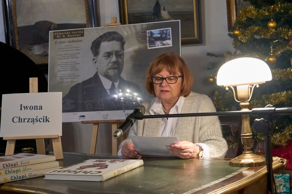 Kobieta okularach siedzi przy zabytkowym biurku, czytając tekst do mikrofonu podczas wydarzenia literackiego. Na stole przed nią znajdują się książki z serii „Chłopi” Władysława Reymonta. Obok stoi tabliczka z jej imieniem i nazwiskiem: „Milena Jastrzębska”. W tle widoczny jest plakat z portretem Władysława Reymonta oraz udekorowana choinka z ozdobami.