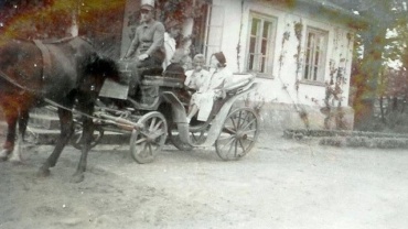 Stare, wyblakłe zdjęcie przedstawiające powóz zaprzężony w konia stojący przed dworkiem z białą elewacją i otwartymi oknami. Na koźle siedzi woźnica w czapce, a w powozie znajdują się starsze kobiety ubrane w jasne stroje. Budynek w tle porośnięty jest bluszczem lub pnączami. Scena ma nostalgiczny, historyczny charakter.