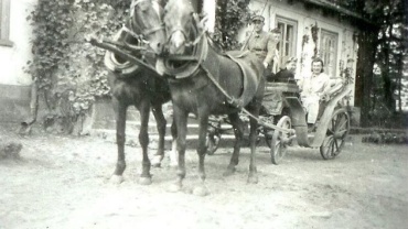 Czarno-białe zdjęcie przedstawiające dwukonny zaprzęg stojący przed dworkiem porośniętym pnączami. Na powozie siedzi woźnica w czapce oraz kilka osób, w tym kobieta ubrana w jasny płaszcz, patrząca w stronę aparatu. Konie stoją na pierwszym planie, a w tle widać budynek z otwartymi oknami i ozdobionymi ścianami.